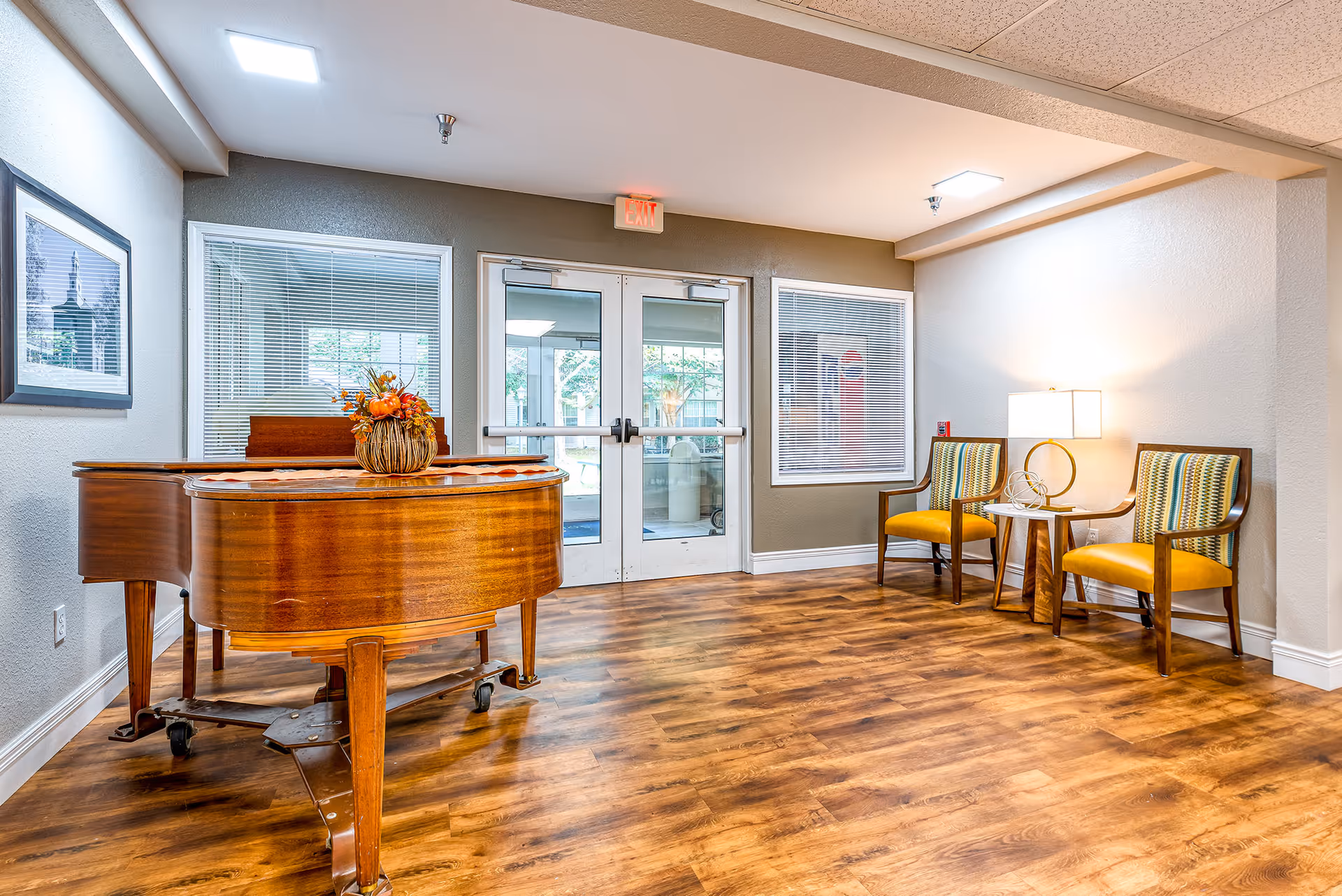 Bright senior living lobby with a wooden grand piano, two yellow chairs by a side table and glass double entrance doors.