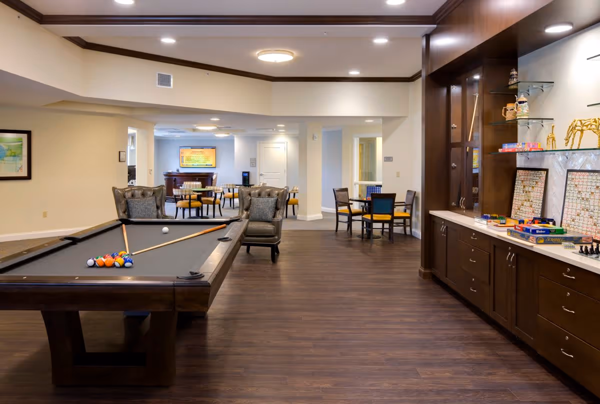 A spacious recreational room featuring a pool table with balls and cues, two armchairs, and several tables with chairs in the background. The room has wooden flooring, recessed ceiling lights, and a built-in cabinet on the right side displaying board games and decorative items.