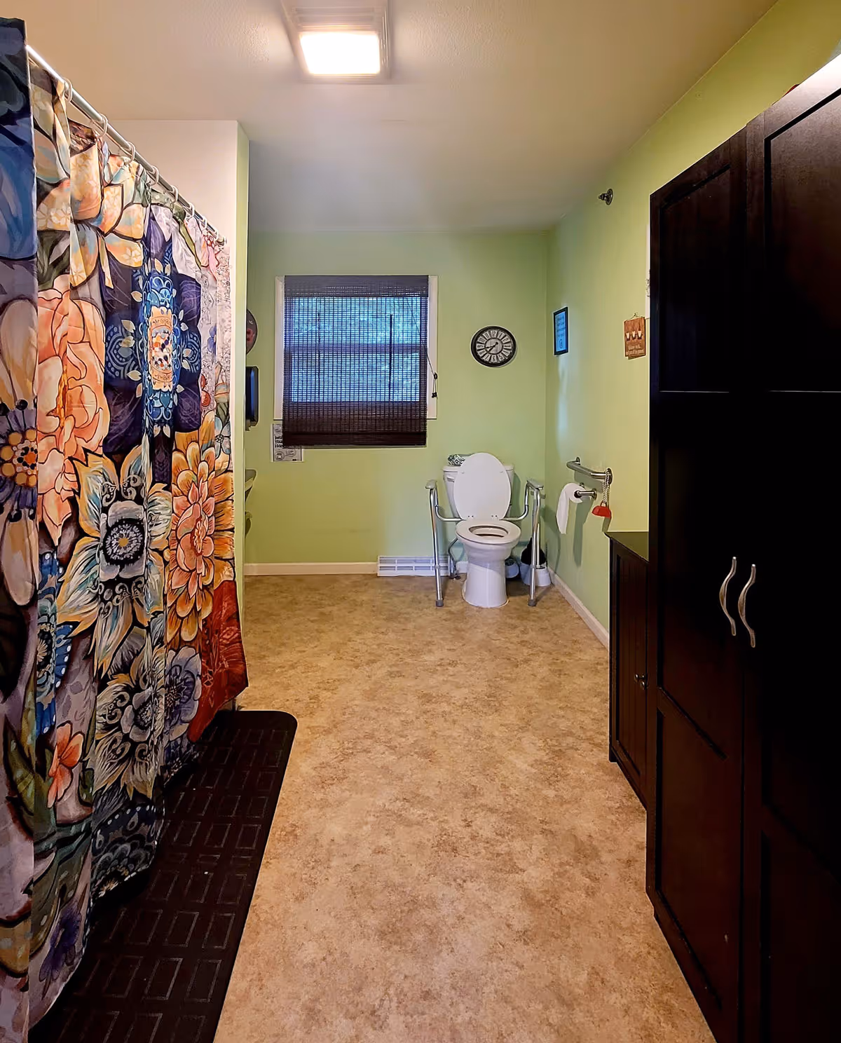 Spacious bathroom with a toilet equipped with grab bars, a colorful floral shower curtain, green walls, and a dark storage cabinet.