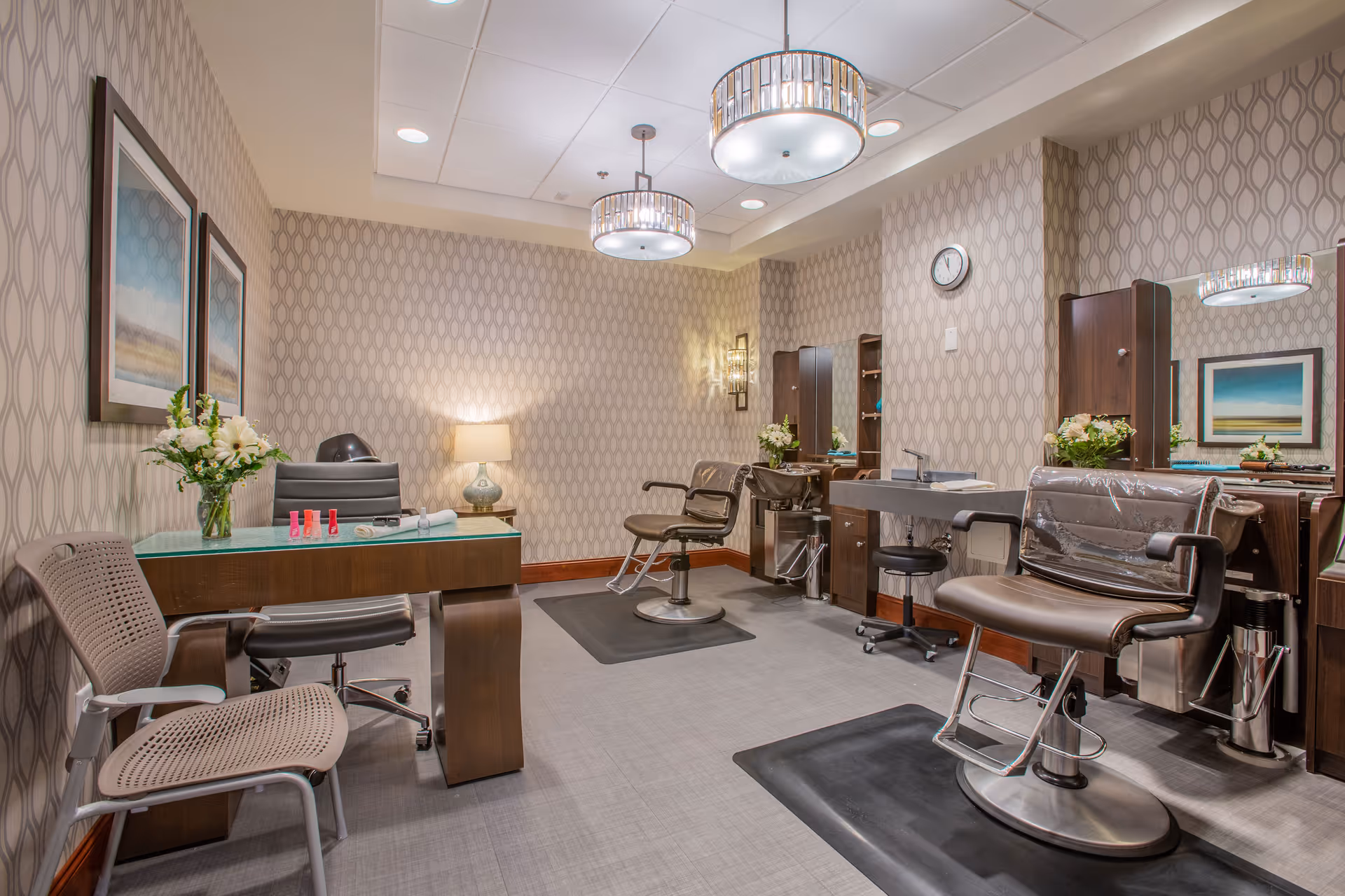 A well-lit salon room with two brown salon chairs on black mats, a manicure table with a chair, and a desk with a black office chair. The walls have a patterned wallpaper, and there are framed pictures, flowers in vases, a clock, and modern ceiling lights.