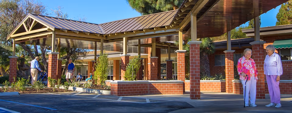 Outdoor covered walkway area with brick pillars and a metal roof at a senior living facility. Several elderly people are standing and sitting nearby, enjoying the sunny day with trees and shrubs around.