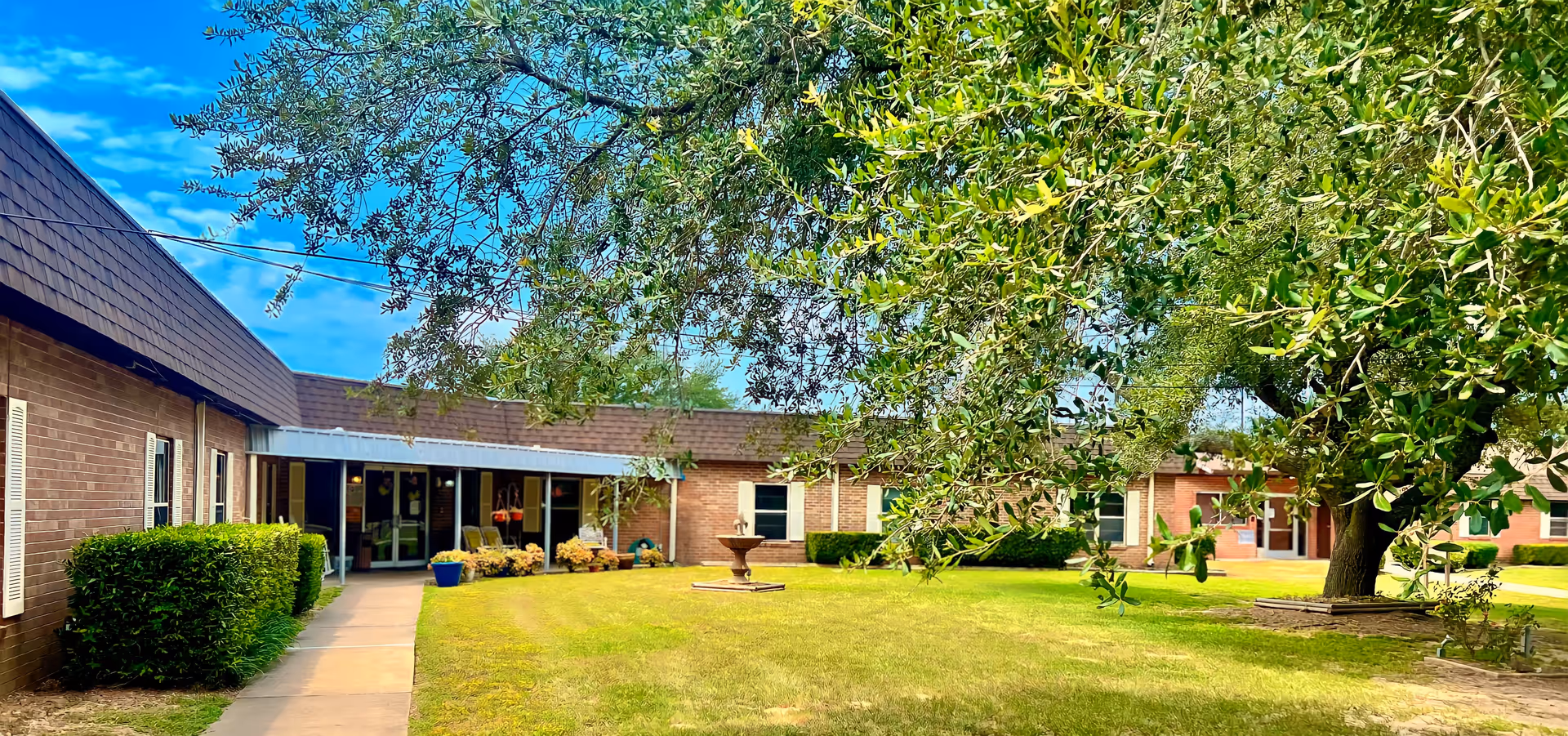 A courtyard area at Capstone Healthcare of Hughes Springs featuring a green lawn, a large tree with spreading branches, a central water fountain, and a surrounding single-story brick building with windows and a covered walkway.