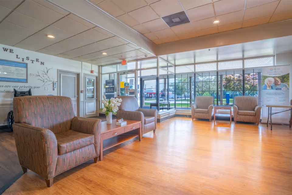 A bright and spacious nursing home lobby with large windows allowing natural light to fill the room. The seating area includes several cushioned armchairs arranged around wooden coffee tables with a vase of flowers and a tissue box. The walls feature a sign that says 'WE LISTEN' and other informational posters. The entrance door is glass, and outside greenery is visible.