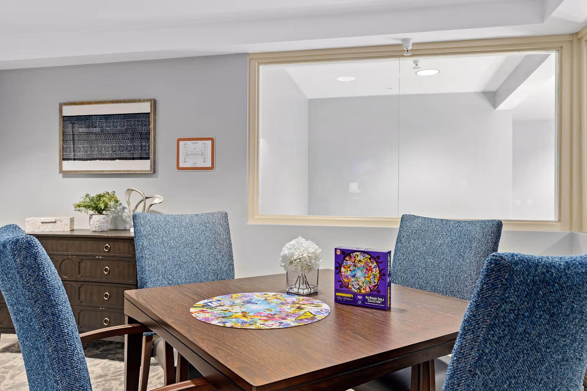 A dining table surrounded by blue upholstered chairs with a colorful puzzle and a small flower arrangement on top, set in a light-gray interior with a sideboard and framed art.