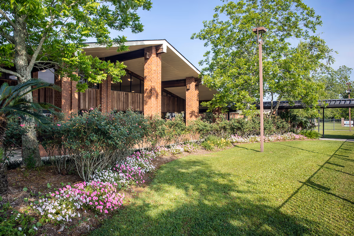 Brick nursing home building front with a covered walkway, manicured lawn, flowering beds, trees, and a lamppost.
