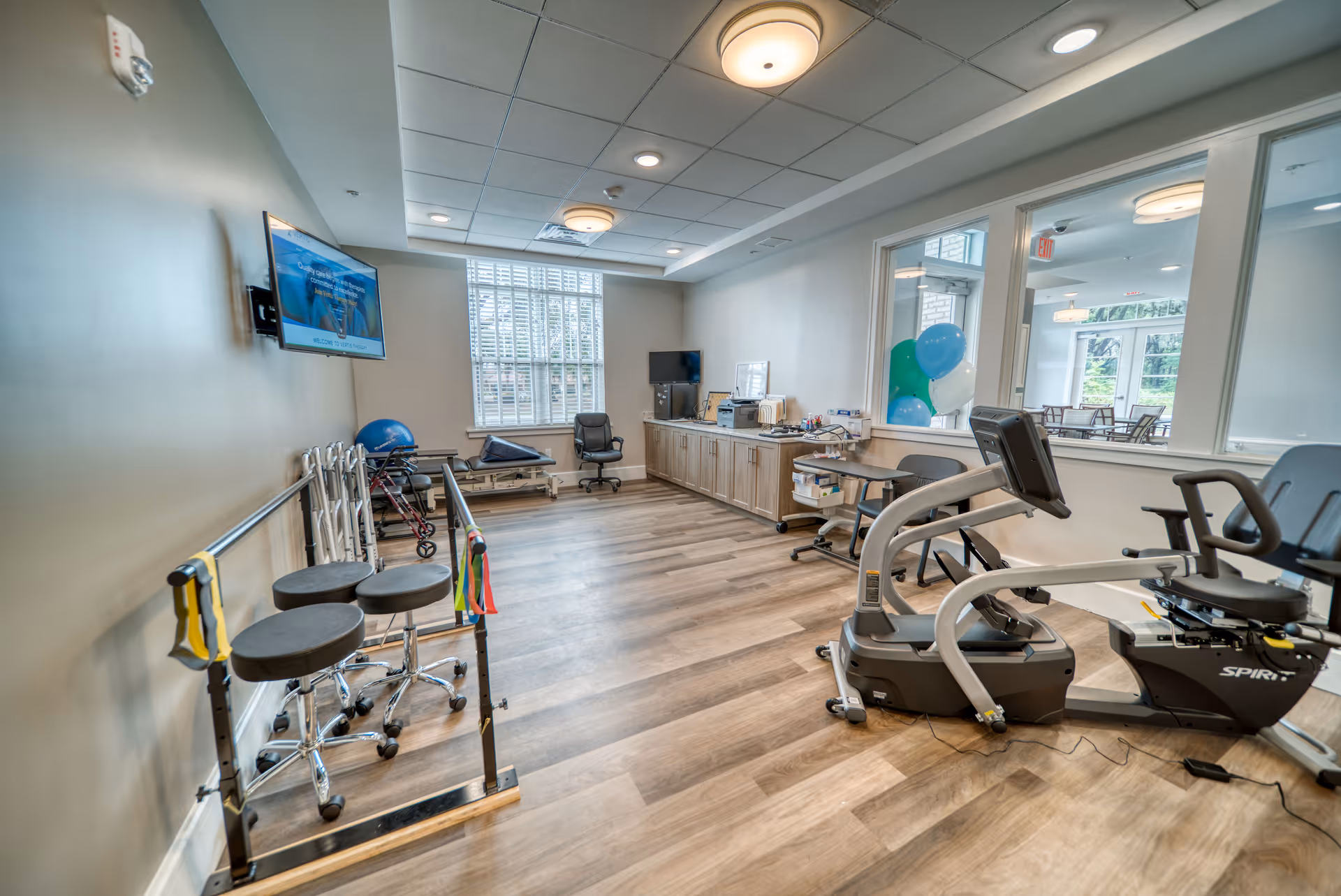 A well-lit physical therapy or rehabilitation room with exercise equipment including a stationary recumbent bike, parallel bars with stools, therapy balls, and a treatment table. The room has wood flooring, a large window with blinds, a wall-mounted TV, and cabinets along the back wall. There is a small desk and chair near a window looking into another room with tables and chairs.