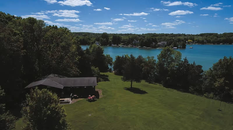 A scenic outdoor view of a green lawn with a small pavilion or shelter on the left side, surrounded by trees. In the background, there is a large blue lake with houses along the shoreline under a partly cloudy sky.