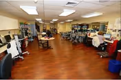 A large room with wooden flooring and fluorescent ceiling lights, containing multiple desks and chairs arranged along the walls and in the center. Various medical equipment and supplies are visible, including wheelchairs and walkers. The room appears to be a clinical or therapy area within a healthcare facility.