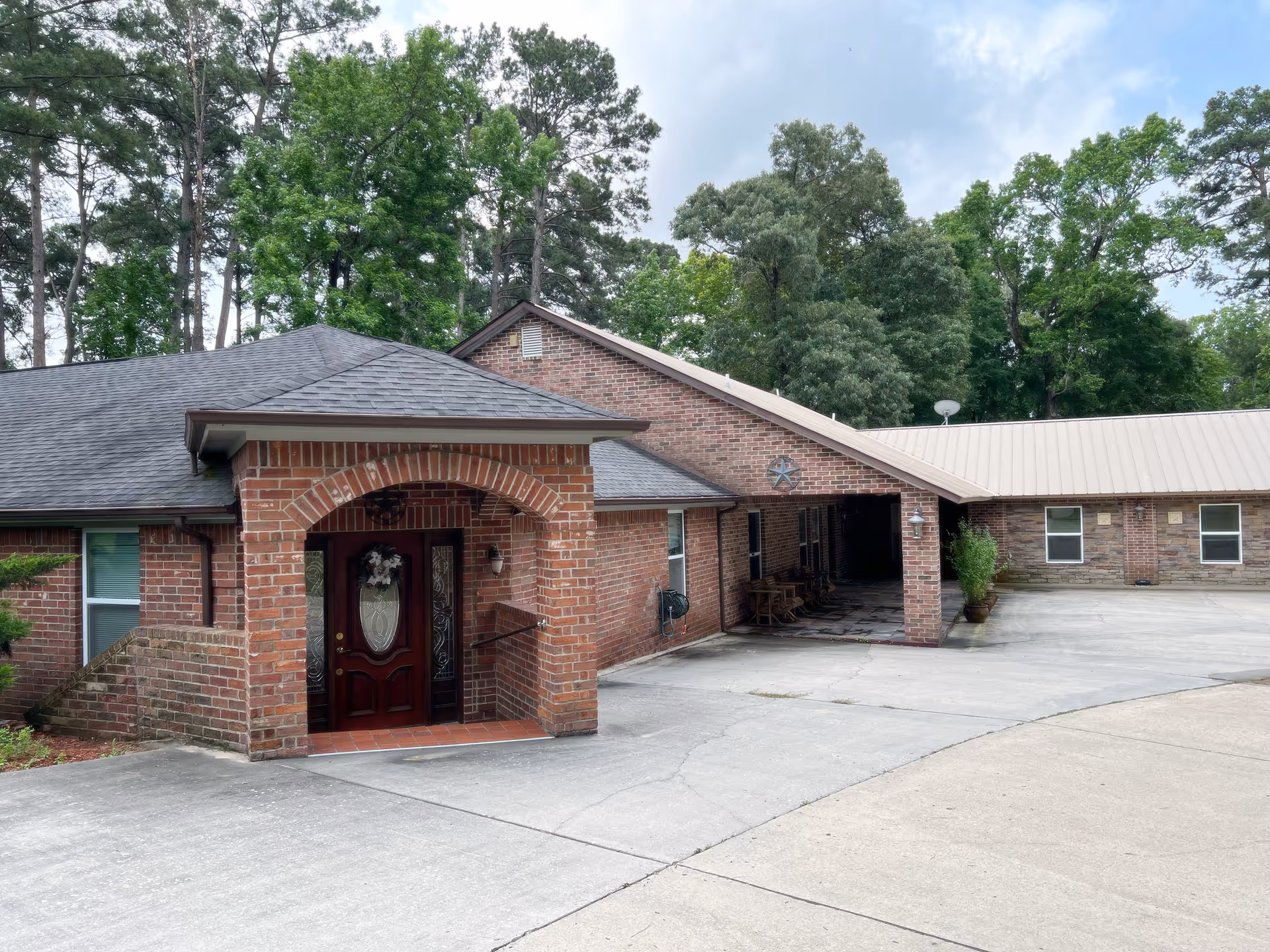 Exterior view of a senior care facility building with red brick walls, a dark shingled roof, and a covered entrance with a decorative glass door. There is a concrete driveway in front and tall green trees in the background under a partly cloudy sky.