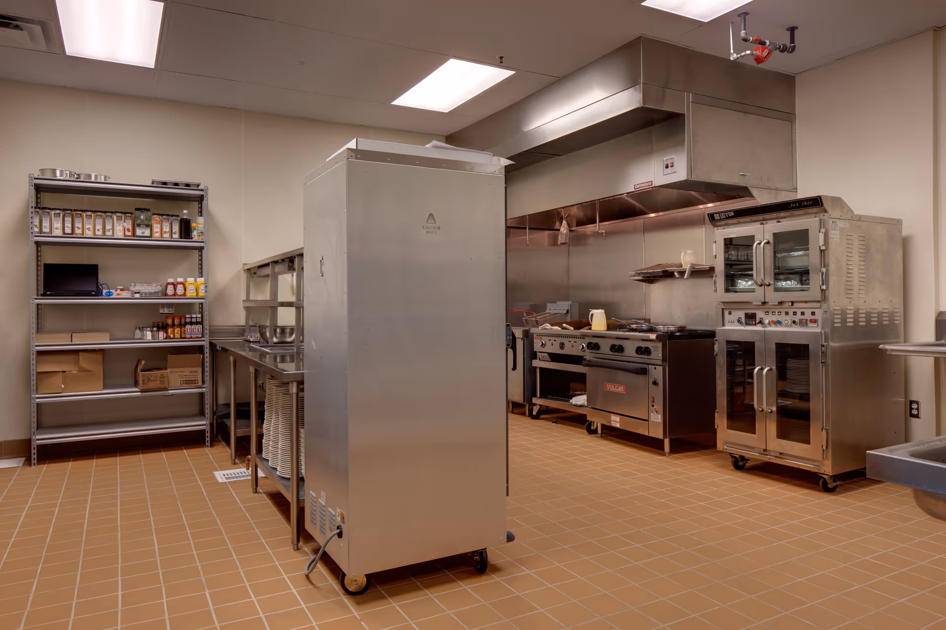 A commercial kitchen with stainless steel appliances, shelving, prep tables, and a tiled floor.