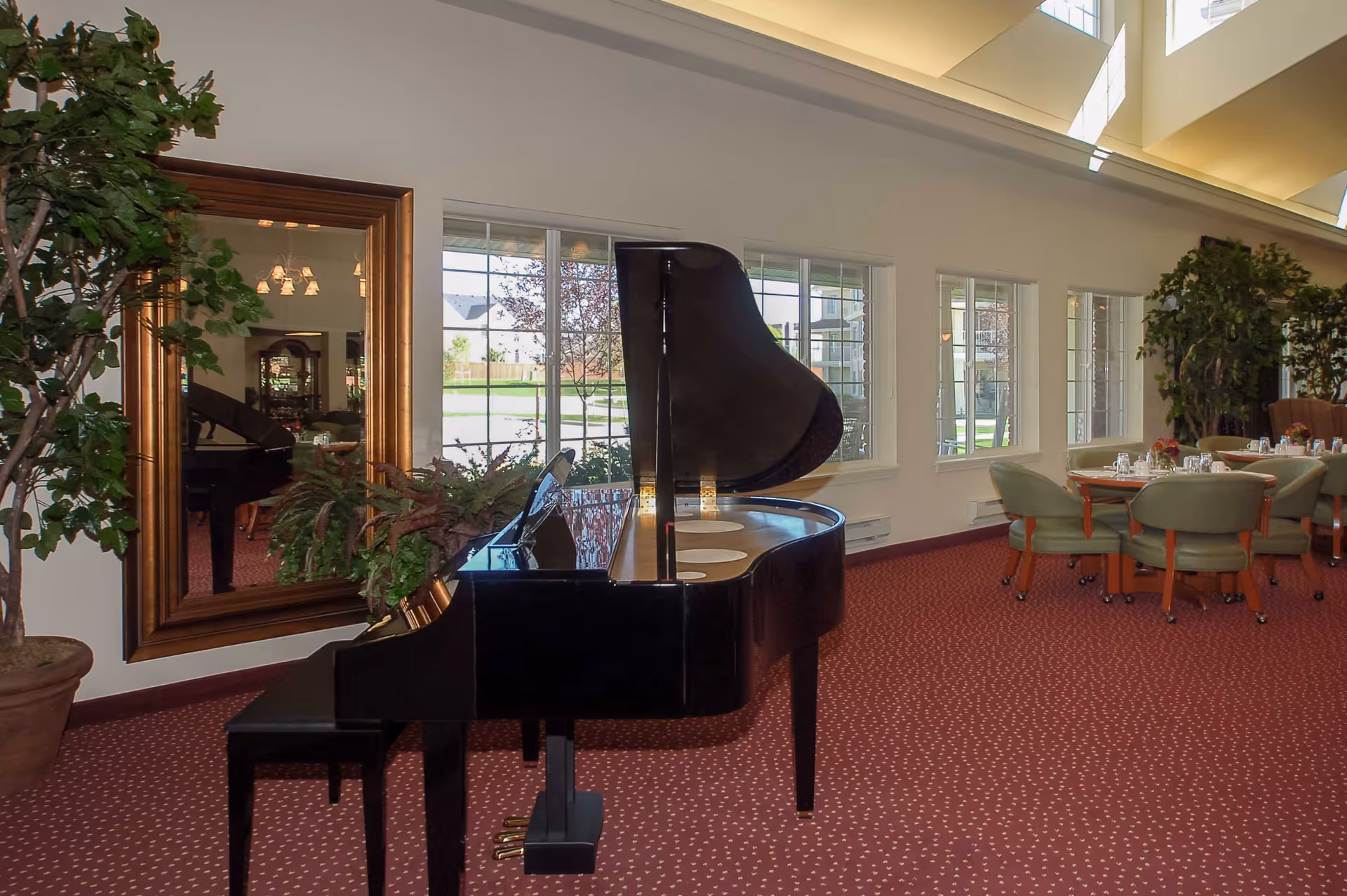 A black grand piano in a carpeted dining room with round dining tables, chairs, large mirror, and potted plants by windows.