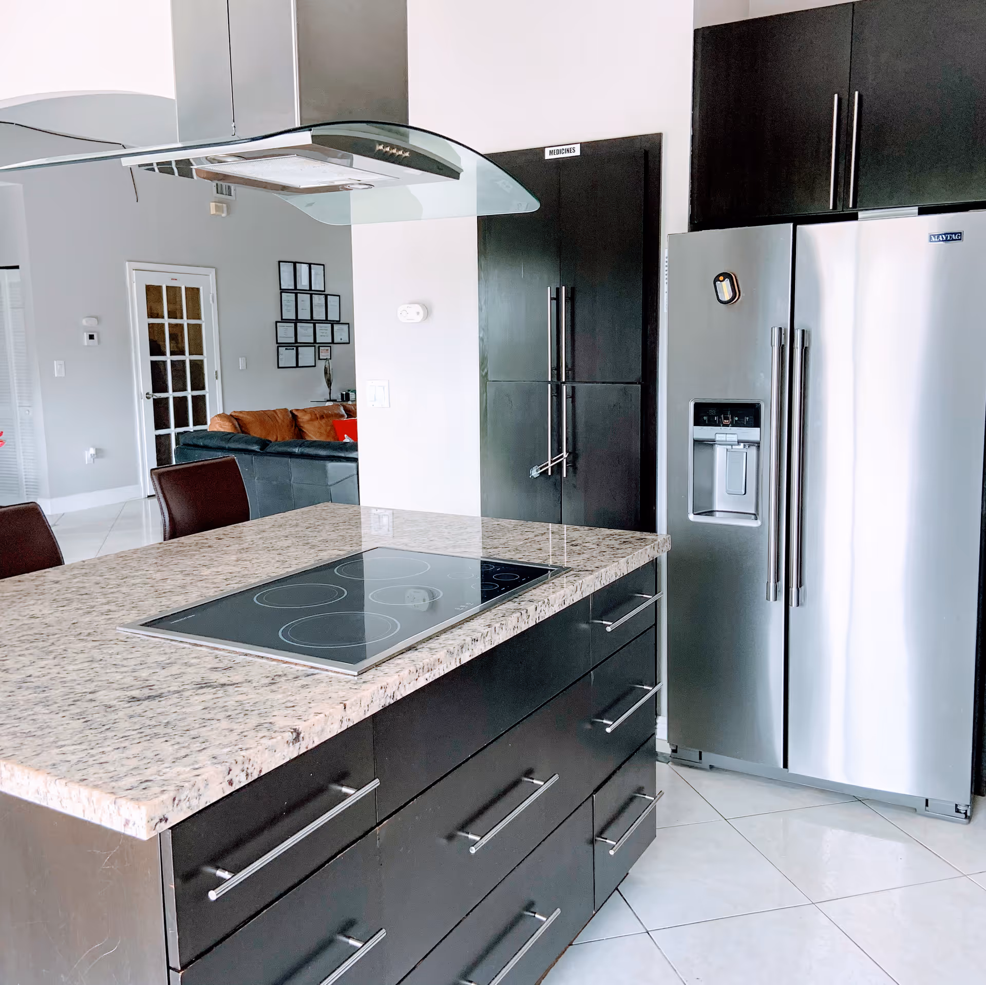 Modern kitchen with a granite countertop island featuring an electric stovetop, dark cabinetry with silver handles, and a stainless steel refrigerator with a water and ice dispenser. In the background, a living area with a brown sofa and wall decorations is partially visible.
