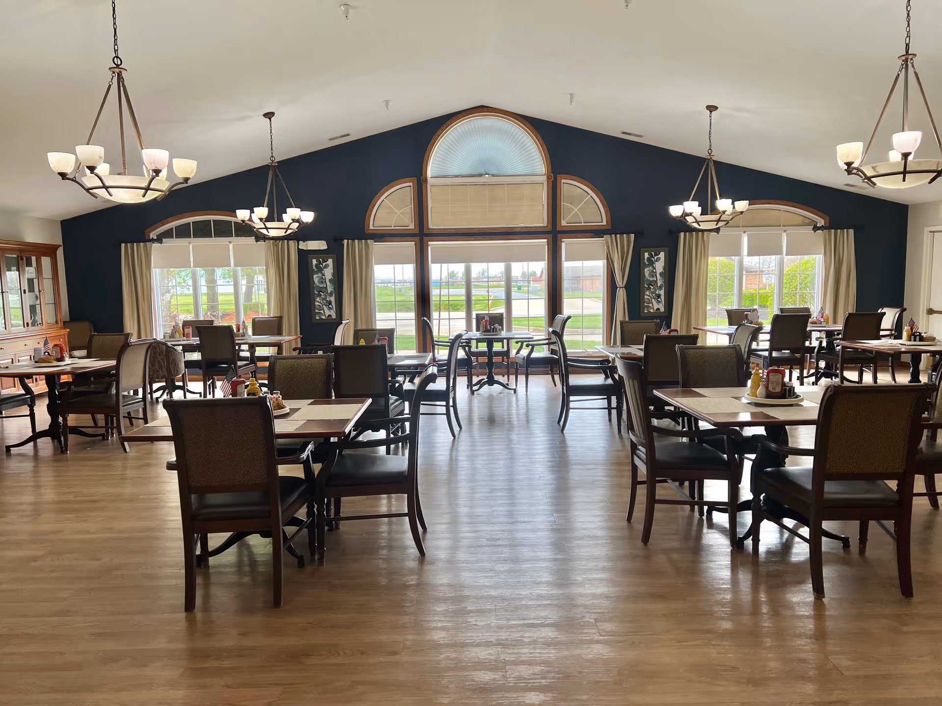 Spacious communal dining room with multiple tables and chairs, chandeliers, and large arched windows overlooking the outdoors.