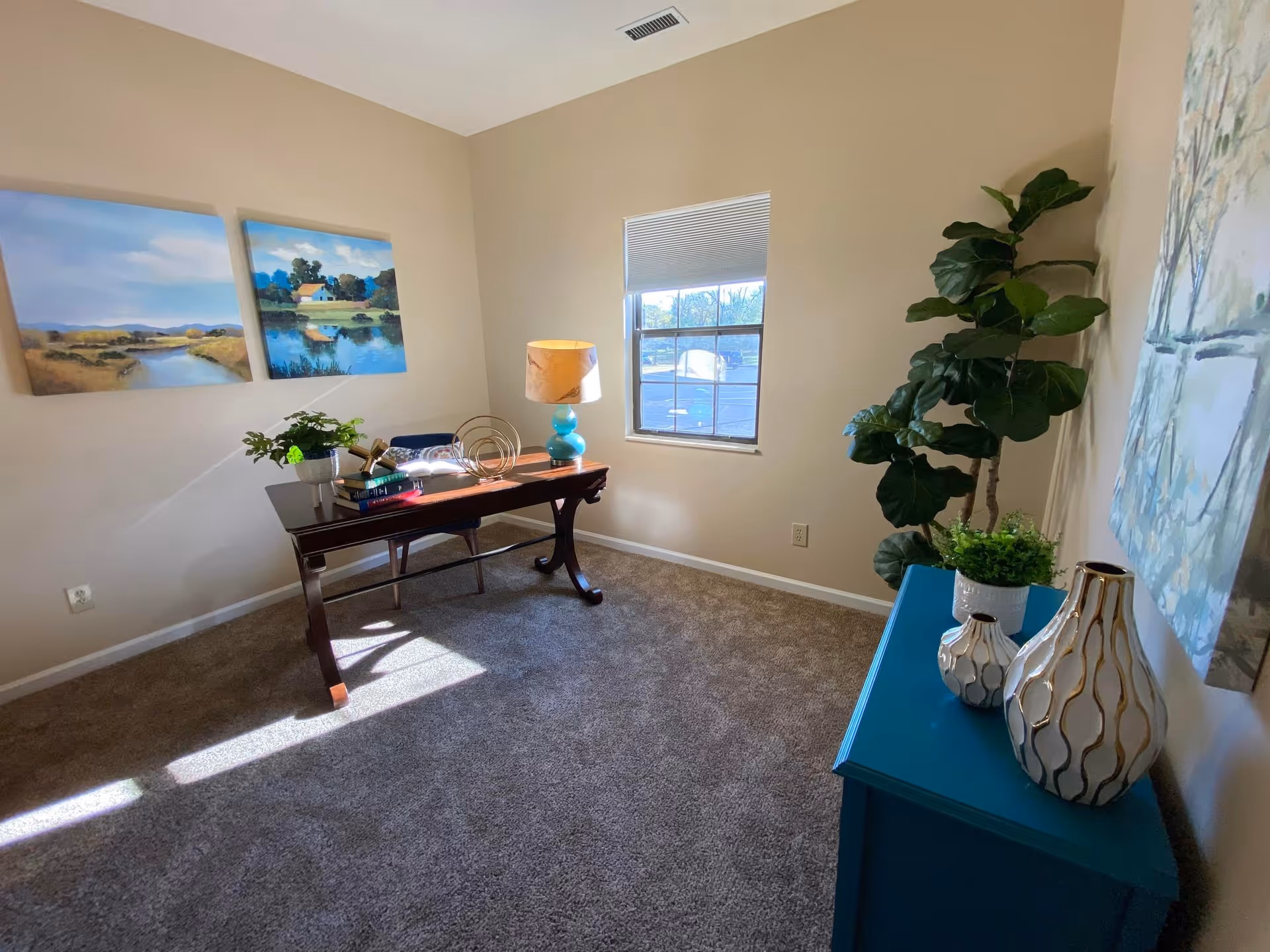 A bright room with beige walls and carpeted floor featuring a wooden desk with books, a lamp, and decorative items. There are two landscape paintings on the left wall, a window with a view outside, a large green plant in the corner, and a blue cabinet with decorative vases and a small plant on top.