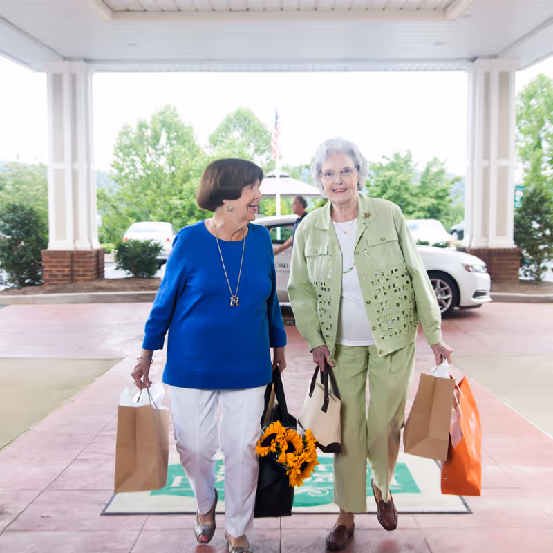 Two elderly women walking outside under a covered entrance, each carrying shopping bags. One woman is wearing a blue top and white pants, and the other is wearing a light green outfit. There are cars and greenery in the background.