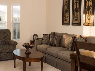 A cozy living room with a sofa, patterned armchair, coffee table, decorative wall art, and shuttered windows.