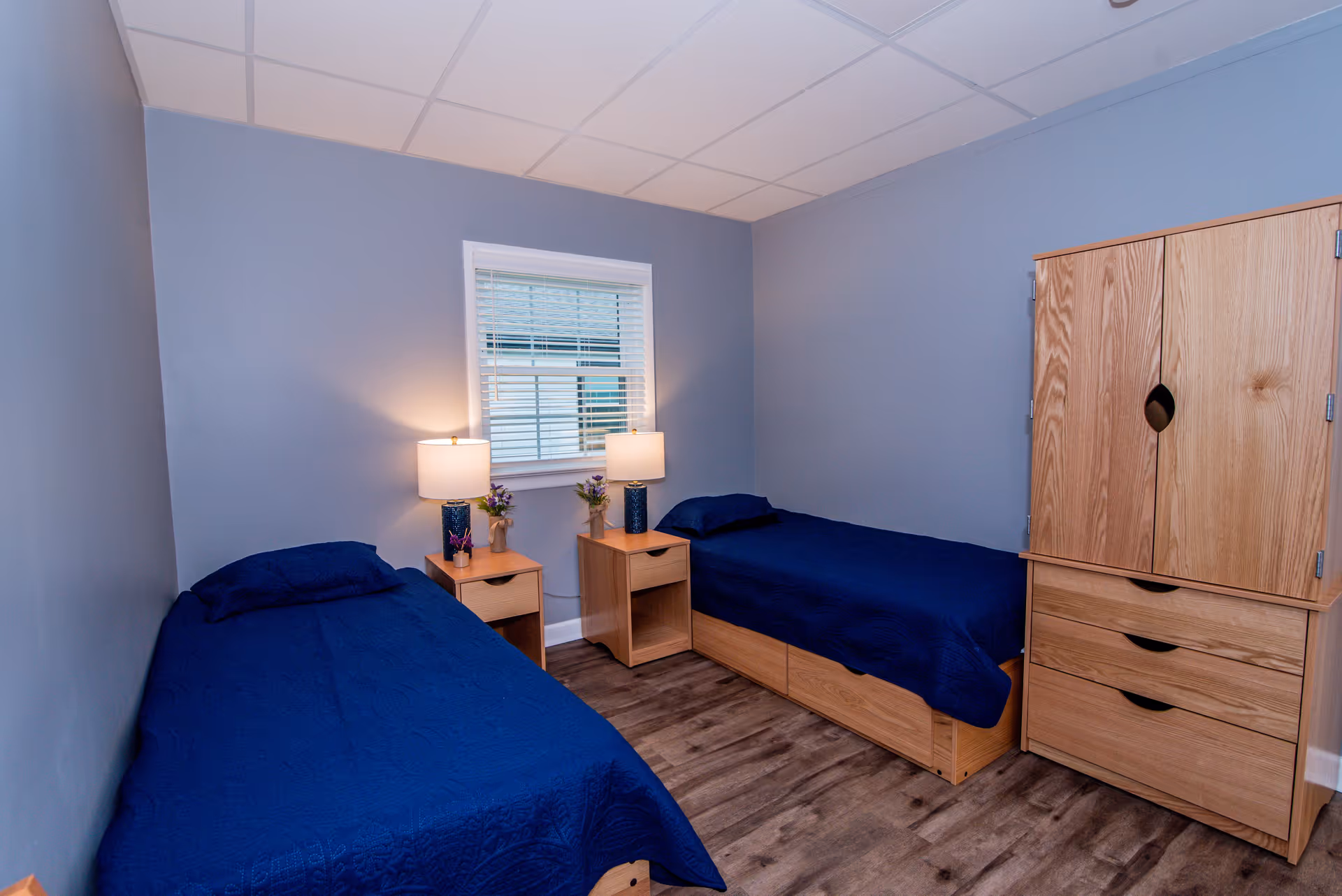 A small bedroom with two single beds covered in dark blue bedding. Each bed has a wooden nightstand beside it with a lamp and small decorative plants. There is a window with white blinds on the wall between the beds. A wooden wardrobe with drawers is positioned against the right wall. The walls are painted light blue and the floor has a wood-like finish.