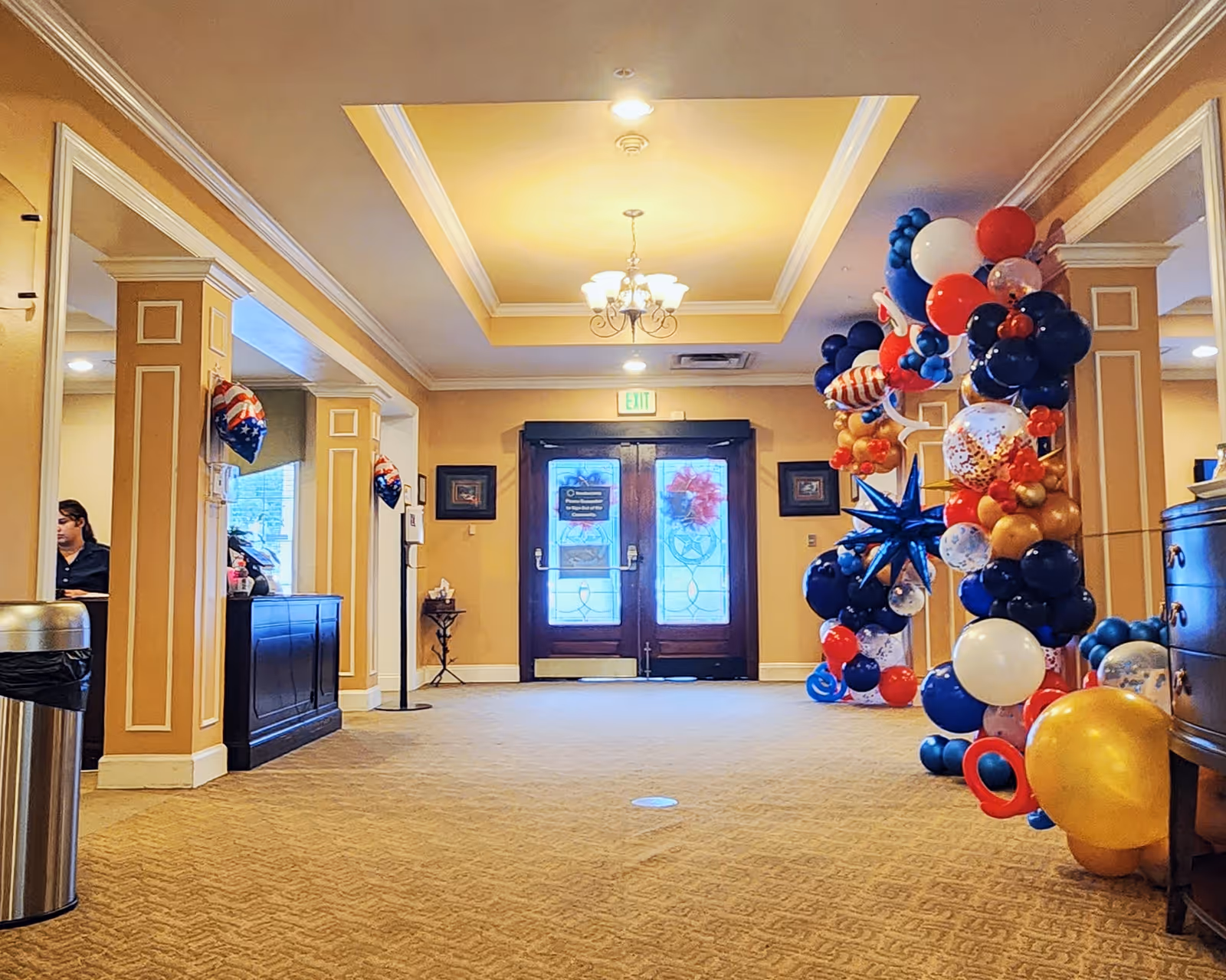 Lobby entrance with double glass doors, chandelier, front desk to the left and large red, white and blue balloon decorations on the right.