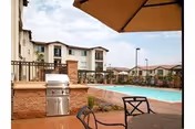 Outdoor patio area with a barbecue grill and a table with chairs under a large umbrella, overlooking a swimming pool and multi-story residential buildings in the background under a partly cloudy sky.