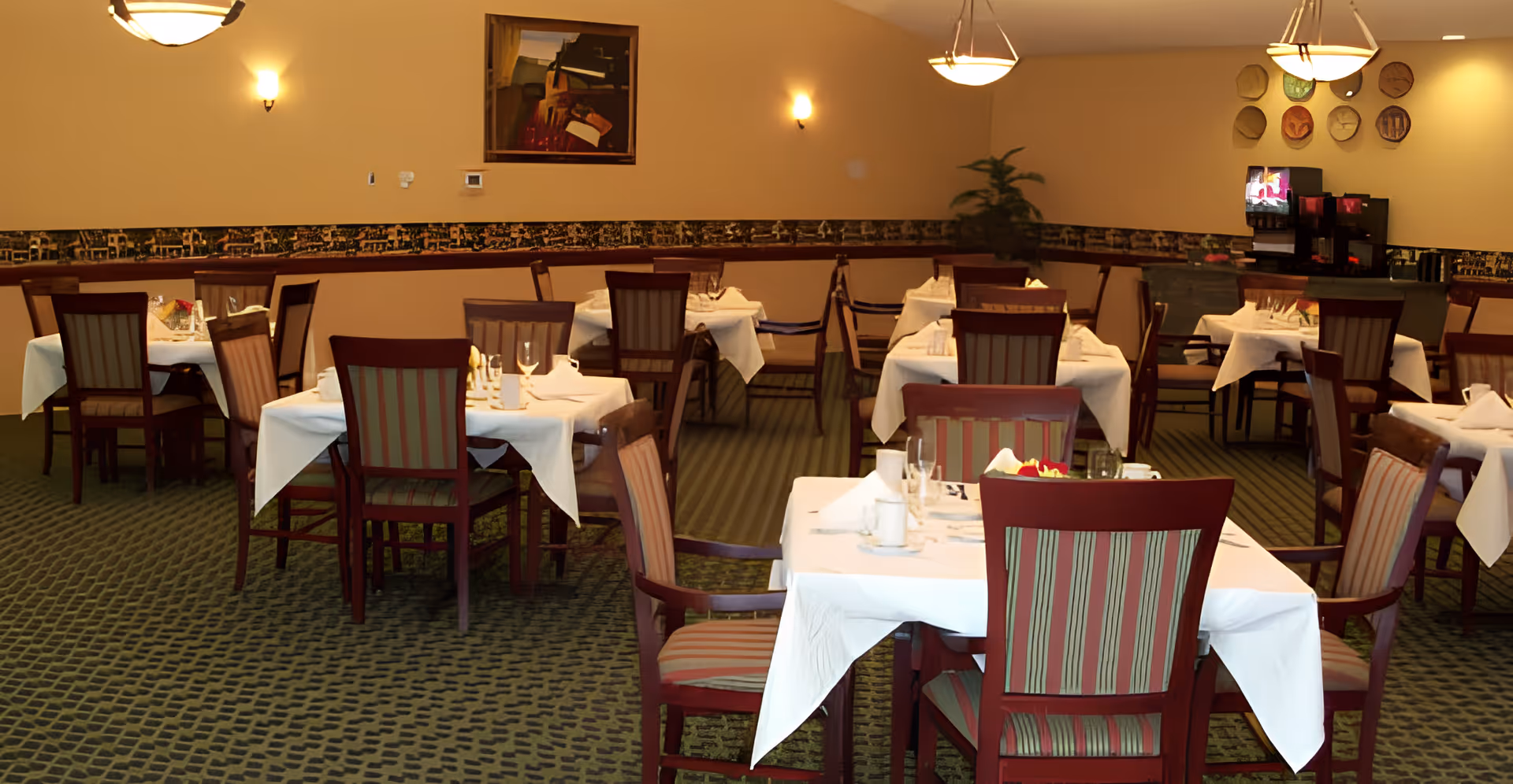 A dining room with multiple tables covered in white tablecloths, each set with glassware and napkins. The room has striped upholstered chairs, carpeted floor, warm lighting from ceiling fixtures and wall sconces, a painting on the wall, and a beverage station in the corner.