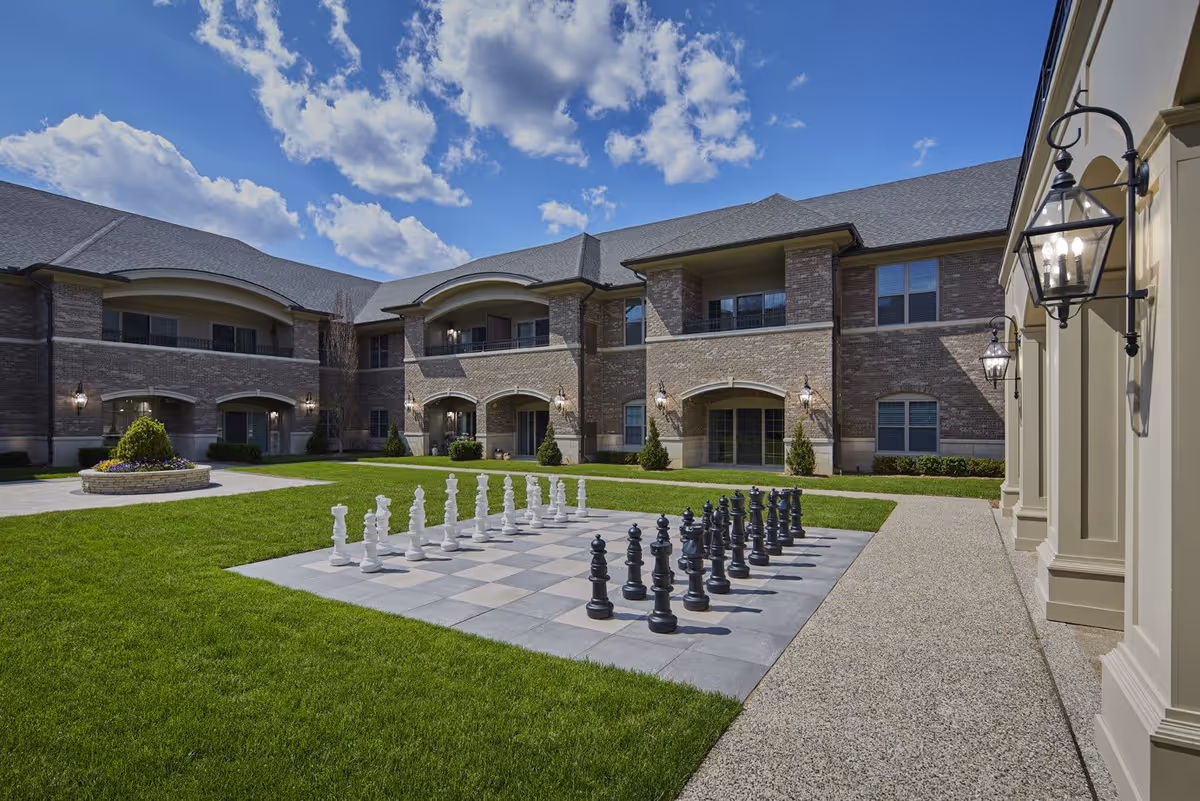 Outdoor courtyard area of a senior living facility with a large chessboard featuring oversized black and white chess pieces on a tiled surface. The courtyard is surrounded by a two-story brick building with balconies and arched doorways. The sky is blue with scattered clouds.