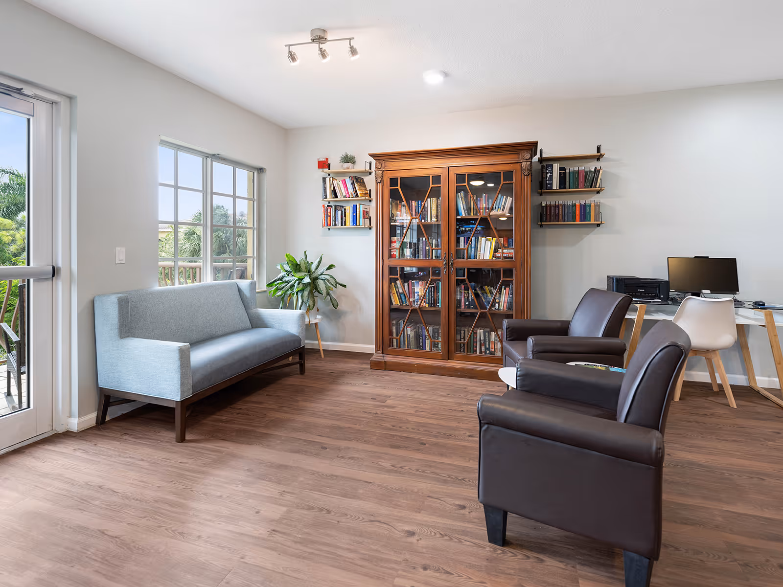 A bright and cozy living room area with a light blue sofa near large windows, two dark brown armchairs facing a small white table, a wooden bookshelf filled with books, and a desk with a computer and printer against the wall.