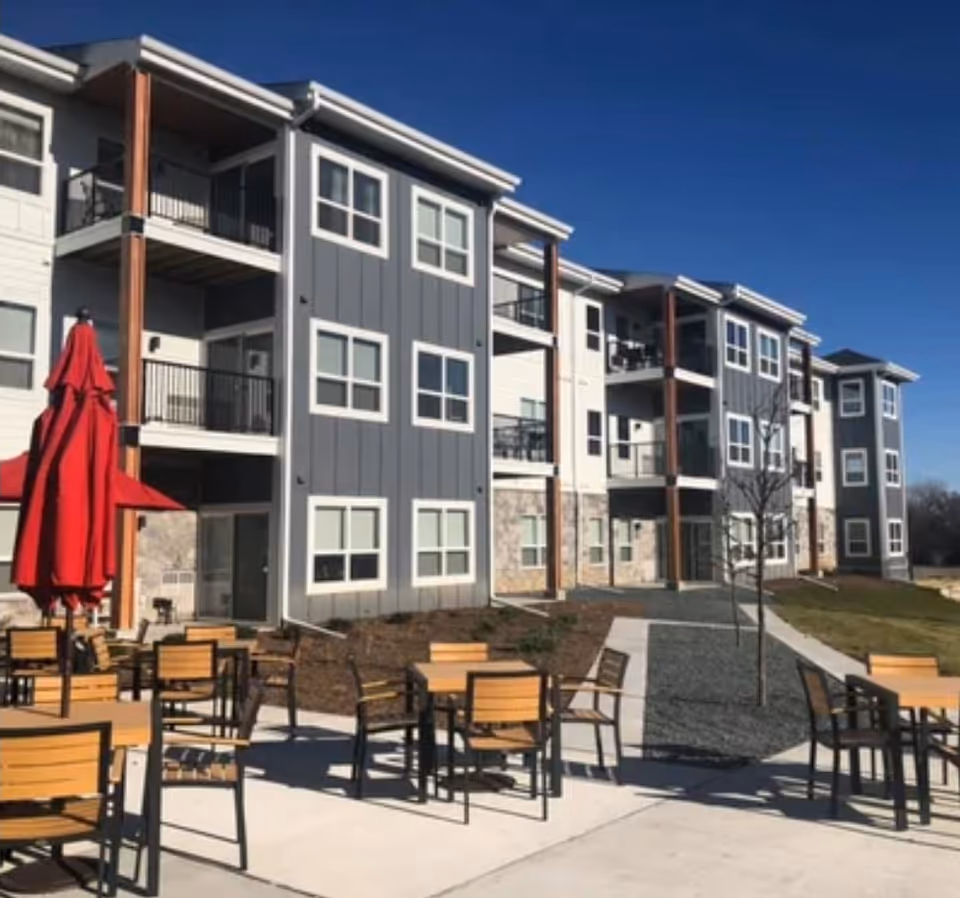 Outdoor patio area with multiple wooden tables and chairs, some with red umbrellas, in front of a modern three-story residential building with balconies and large windows under a clear blue sky.