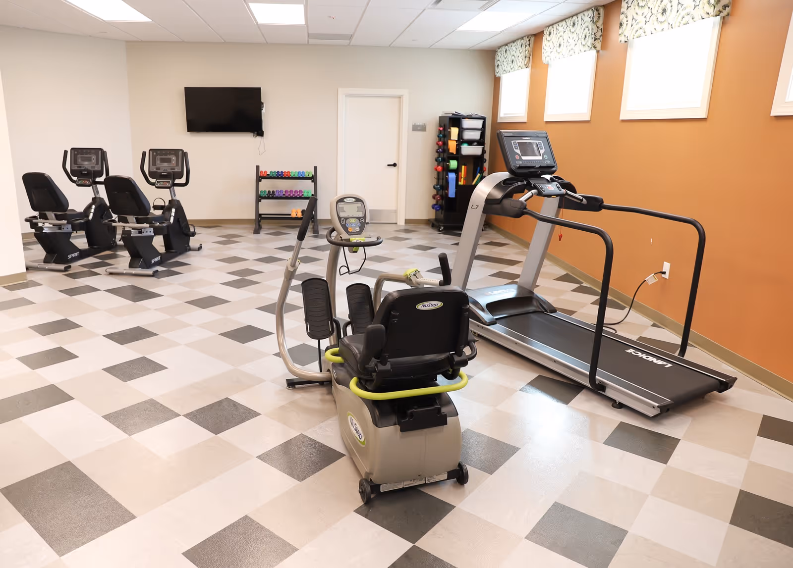 A fitness room with exercise equipment including a treadmill, two recumbent bikes, and a stationary elliptical machine. There is a rack with colorful dumbbells and a wall-mounted TV. The room has a checkered tile floor, beige and orange walls, and windows with patterned valances.