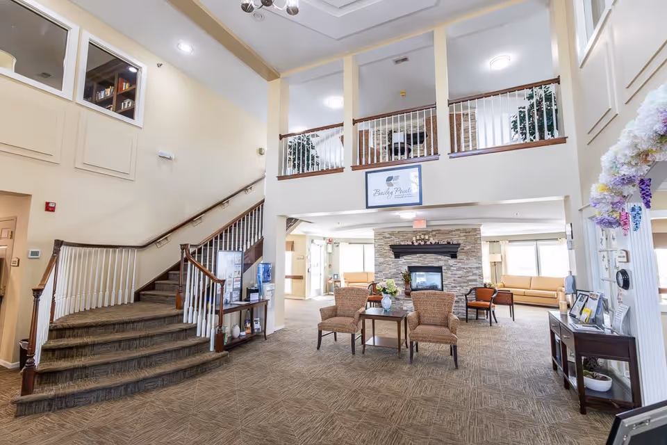 Spacious senior living facility common area with a carpeted floor, a staircase with wooden handrails leading to an upper level, a stone fireplace with a mantle, several chairs and tables arranged for seating, large windows allowing natural light, and a sign above the fireplace that reads 'Bailey Pointe'.