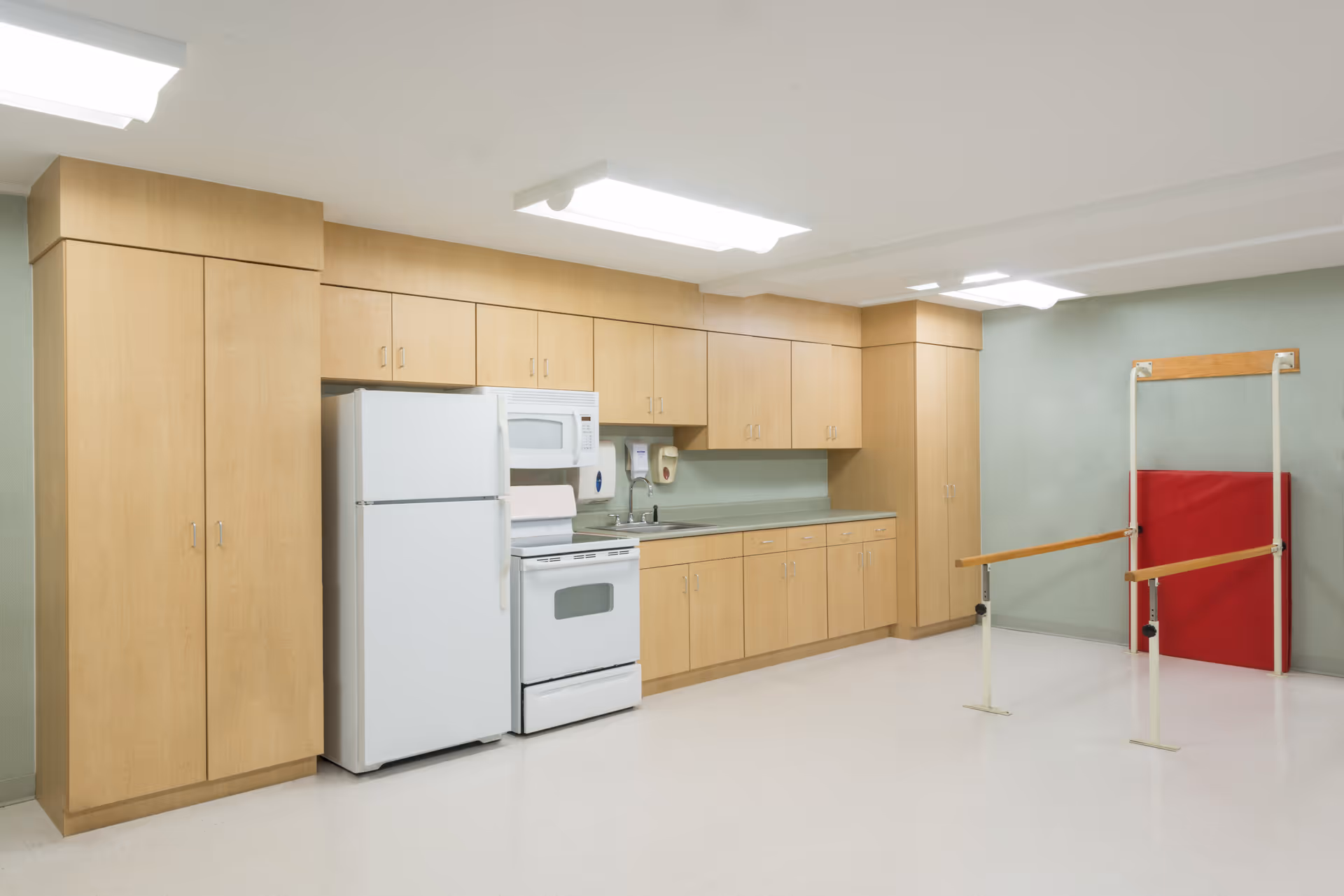 A bright kitchenette with light wood cabinets, a white refrigerator and stove, and parallel bars with a red mat against a pale green wall.