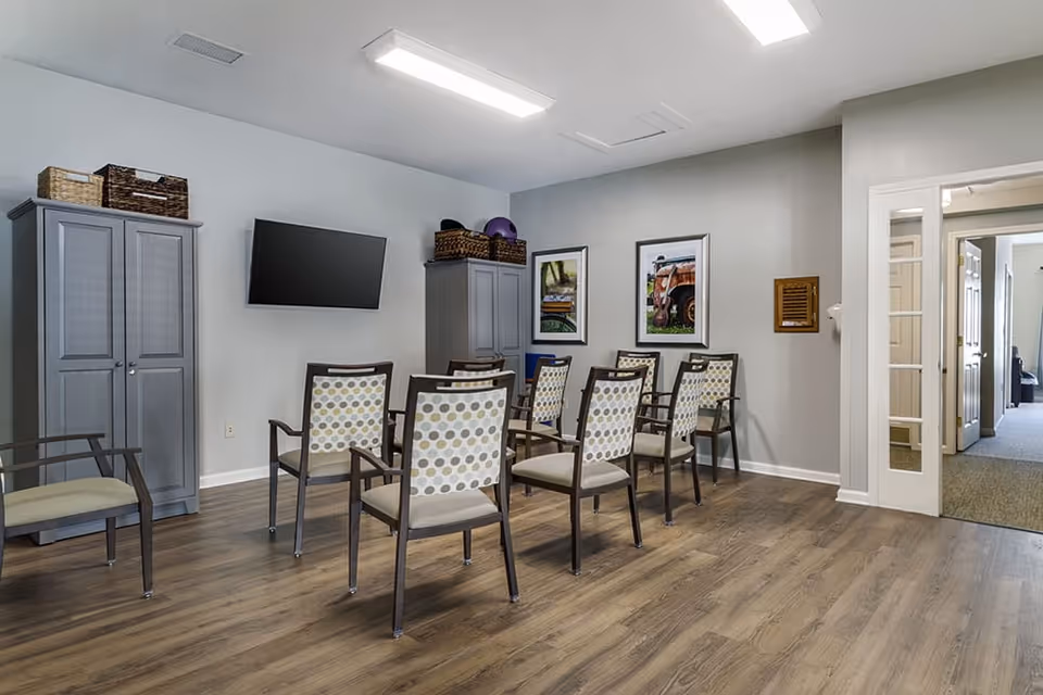 A small meeting or activity room with eight chairs arranged in two rows facing a wall-mounted flat screen TV. The room has light gray walls, wood flooring, two gray storage cabinets with baskets on top, and two framed pictures on the wall. There is a doorway leading to another room with carpeted flooring.