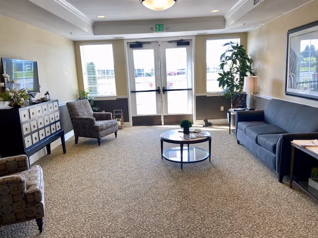 A well-lit senior living facility common area with a patterned carpet, a gray sofa, two patterned armchairs, a round glass coffee table with a small plant, a side table with a lamp, a decorative cabinet with flowers, and double glass doors leading outside.