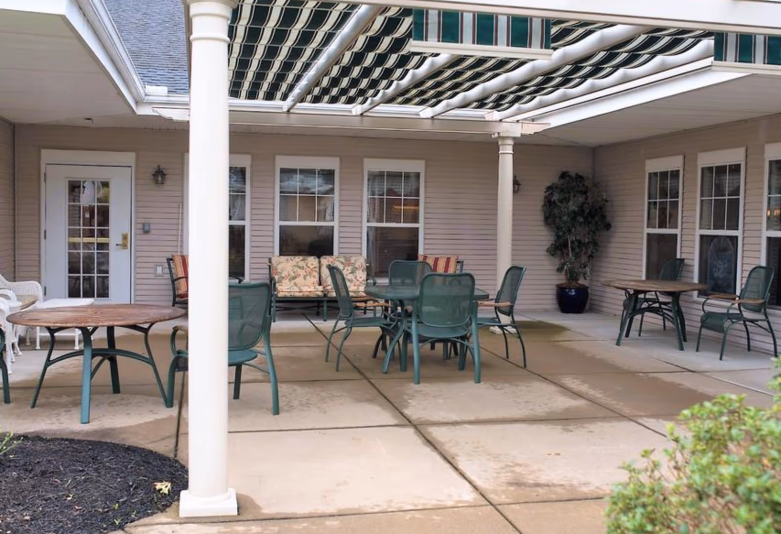 Covered outdoor patio with round tables, green metal chairs, a cushioned bench and supporting columns under a striped awning.