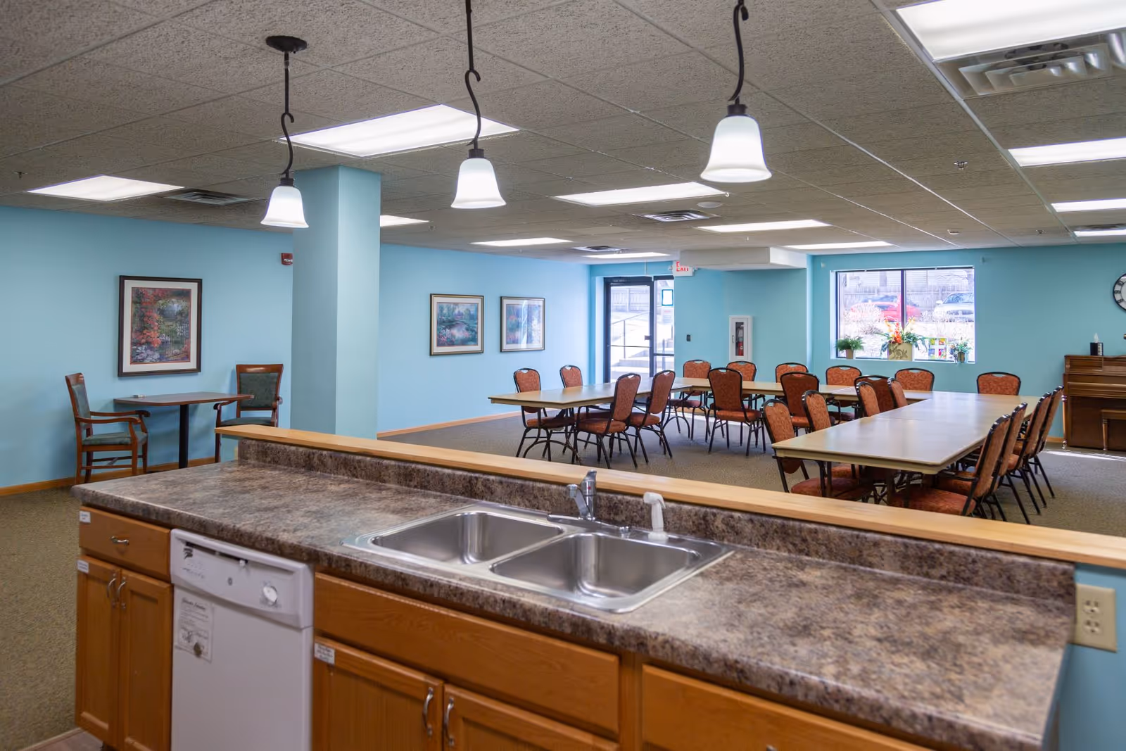 Interior view of a senior living facility dining area with multiple tables and chairs arranged for group seating. The room has light blue walls, framed artwork, and large windows letting in natural light. In the foreground, there is a kitchen counter with a double sink and a dishwasher underneath. Pendant lights hang from the ceiling above the counter.