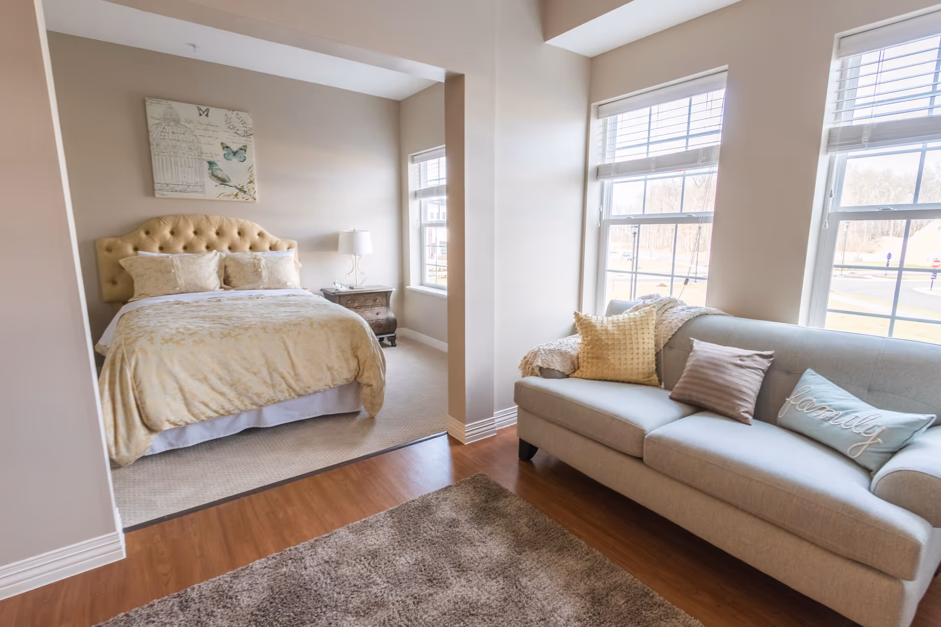 A cozy living space featuring a beige tufted bed with matching bedding and pillows, a wooden nightstand with a lamp, and a light gray sofa adorned with decorative pillows including one that says 'family'. The room has large windows letting in natural light and a soft brown rug on the wooden floor.