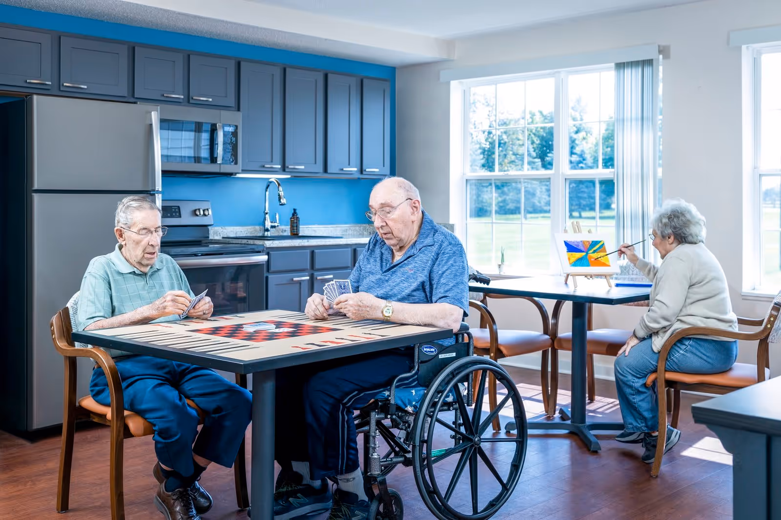 Two elderly men sitting at a table playing cards in a room with a kitchen area in the background. One man is in a wheelchair. Nearby, an elderly woman is seated at another table painting on a small canvas. Large windows let in natural light and show a green outdoor view.