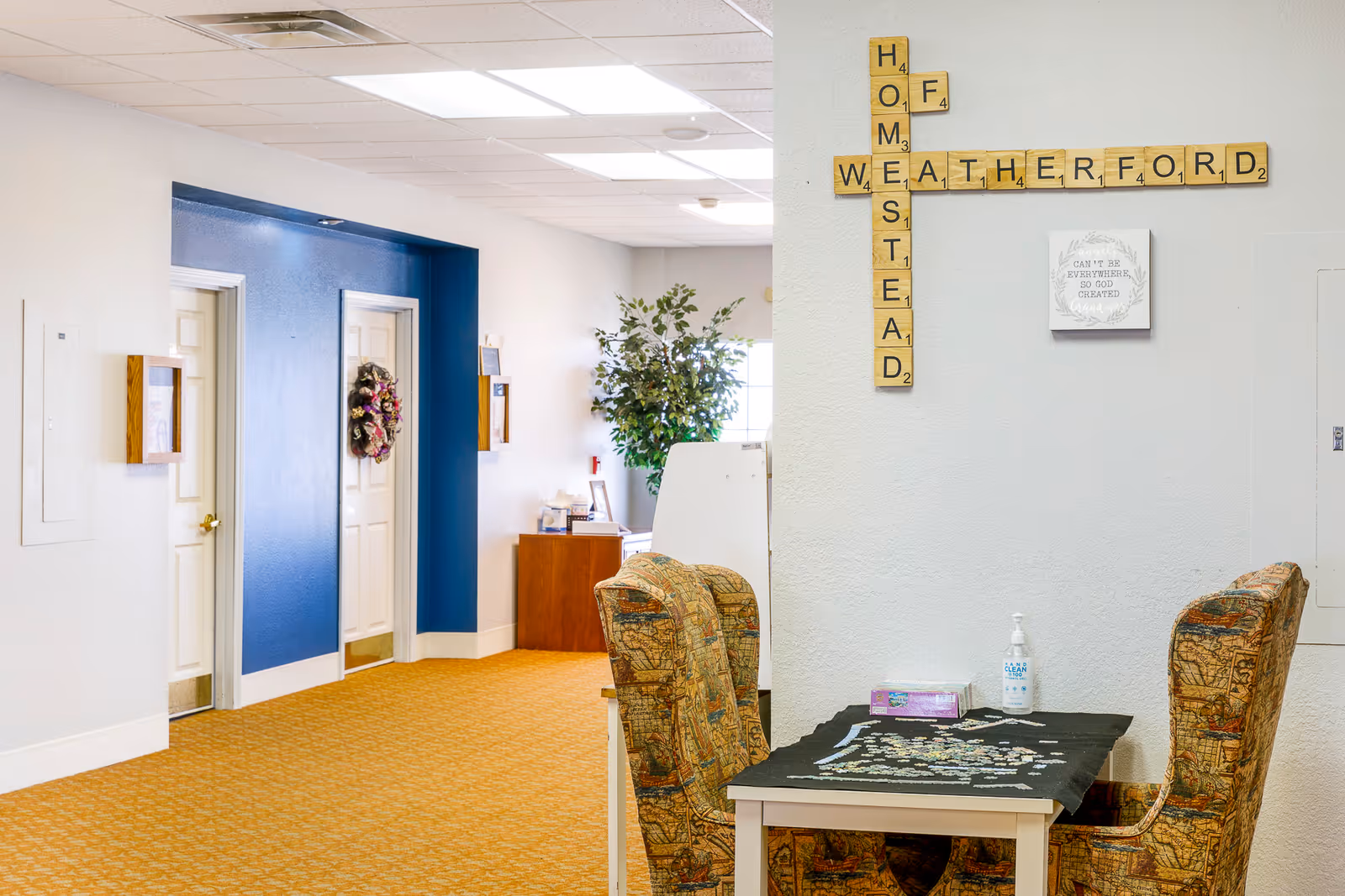 Interior view of a senior living facility hallway with two closed white doors on the left, one decorated with a floral wreath. A small table with two patterned chairs is in the foreground, holding a puzzle in progress, a box of tissues, and a bottle of hand sanitizer. On the wall above the table, large Scrabble-style letter tiles spell out 'HOMESTEAD' vertically and 'WEATHERFORD' horizontally. A framed sign with a quote is also visible on the wall. The hallway has a blue accent wall and a potted plant near a window in the background.