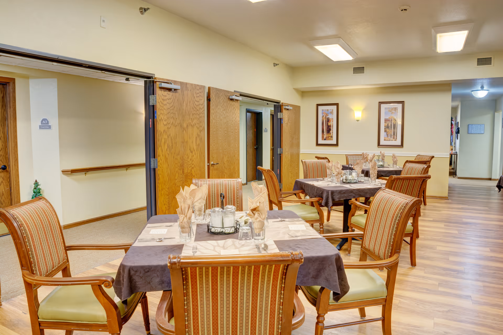 Interior view of a dining room in an assisted living facility with tables set for meals. The tables have tablecloths, napkins, glasses, and condiments. There are wooden chairs with cushioned seats around the tables. The room has wooden flooring, light-colored walls, framed artwork, and open double doors leading to a hallway.