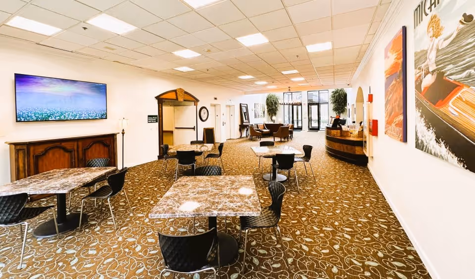 Bright communal dining and lounge area with multiple small marble-top tables and chairs, a reception desk, and wall art.