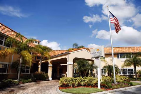 Heron House Assisted Living entrance with tropical landscaping, a flagpole, and a bright sunny day.
