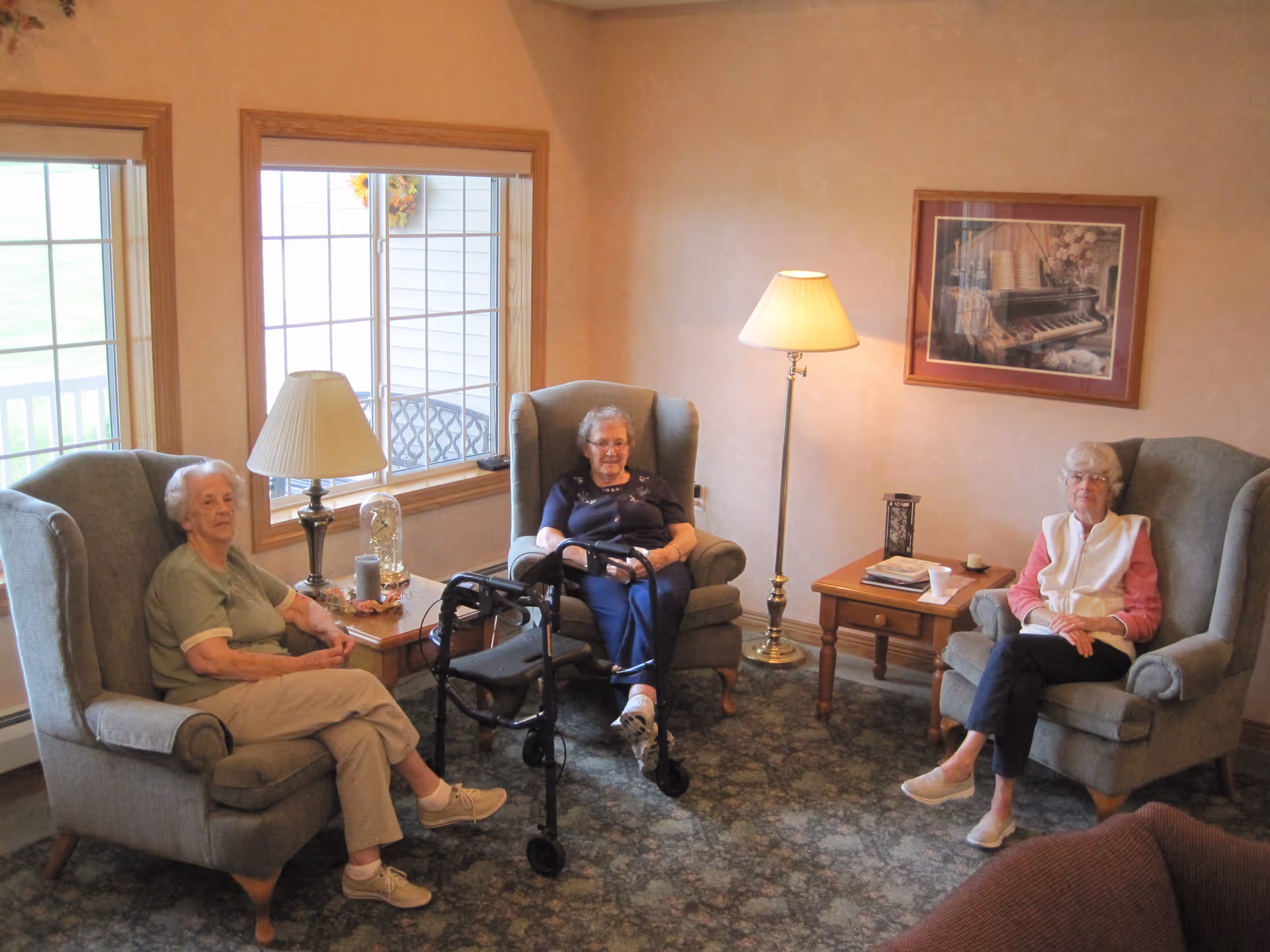 Three elderly women sitting in armchairs in a cozy lounge area with lamps, side tables, and windows.