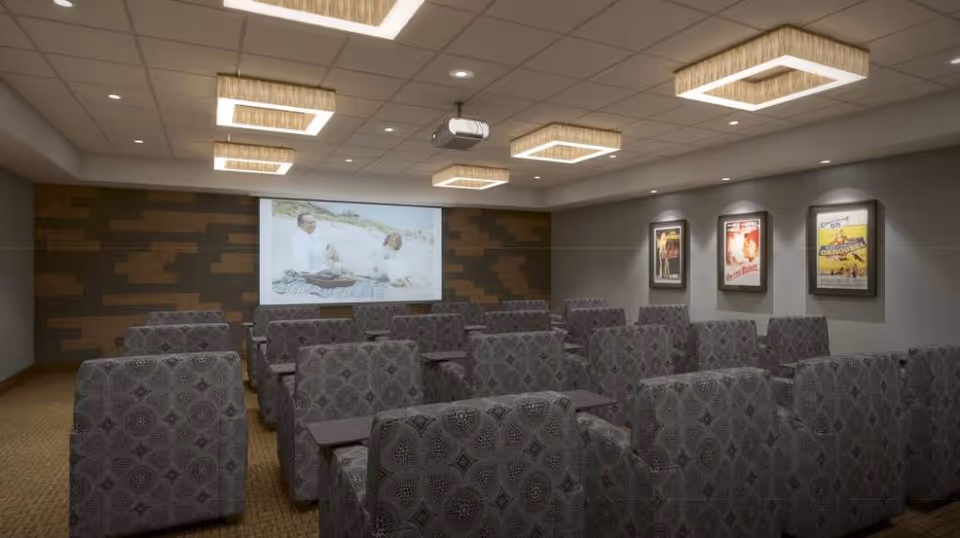 A small theater room with rows of patterned upholstered chairs facing a large screen showing a movie scene. The room has modern square ceiling lights and movie posters on the right wall.