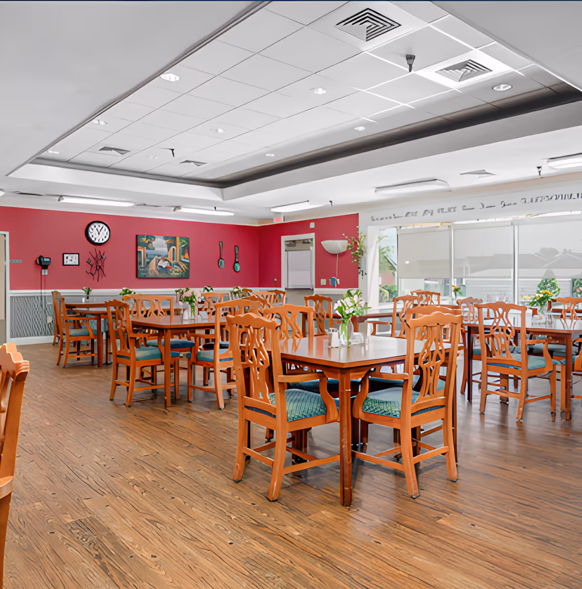 Bright dining room with wooden tables and chairs, vases of flowers on the tables, large windows, and a red accent wall.