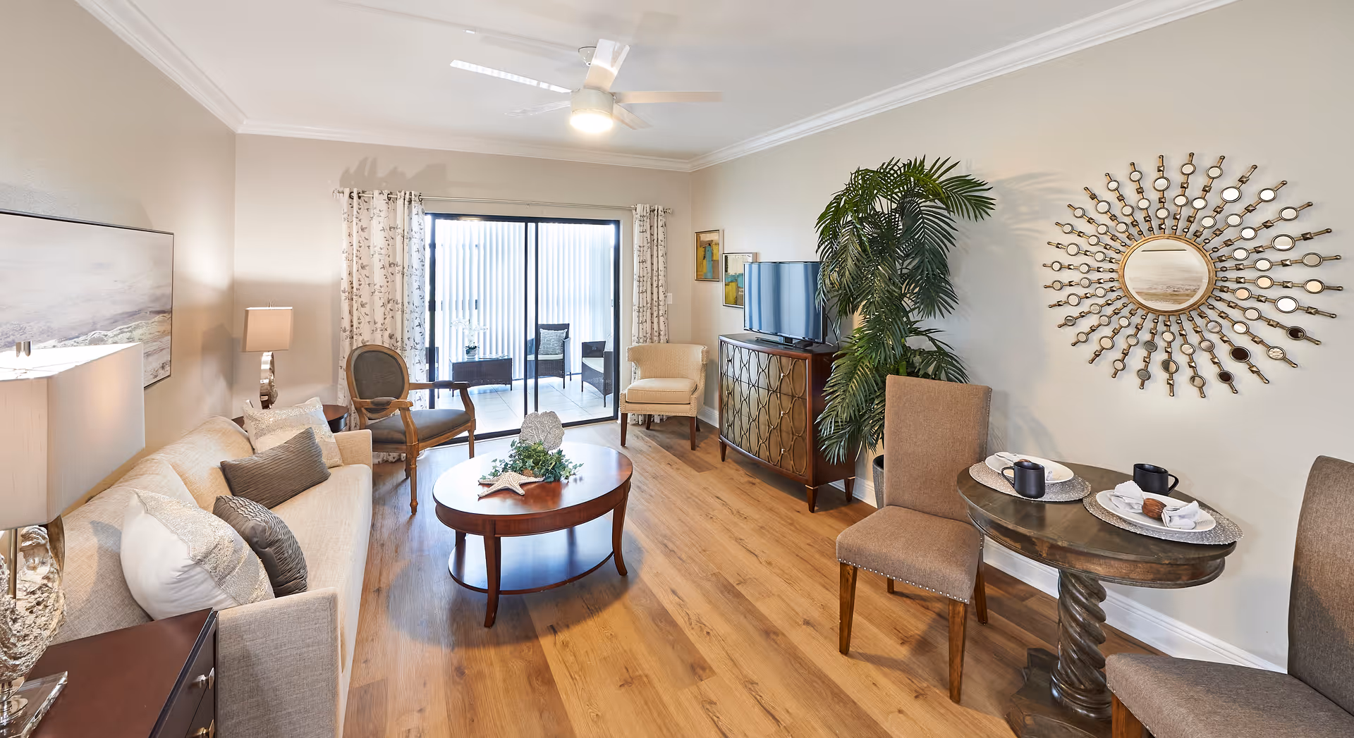 A bright and cozy living room featuring a beige sofa with multiple cushions, a wooden coffee table with decorative items, two armchairs, a TV on a wooden cabinet, a large decorative mirror on the wall, and a small round dining table with two chairs set with plates and mugs. There is a ceiling fan above and sliding glass doors leading to a patio with outdoor seating.
