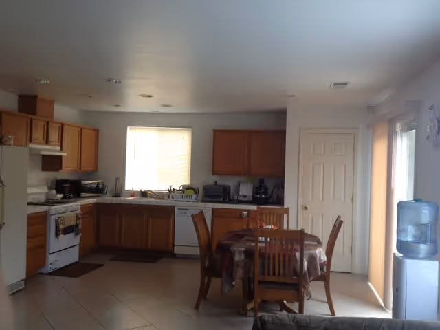 A kitchen and dining area with wooden cabinets, a white refrigerator, stove, and dishwasher. A small dining table with four wooden chairs is covered with a patterned tablecloth. There is a water dispenser near a sliding glass door with vertical blinds, and a window above the kitchen sink letting in natural light.