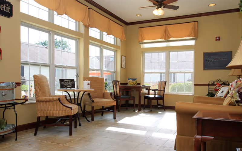 A bright and cozy common area with large windows letting in natural light. The room features cushioned armchairs, a small round table with a sign about coffee, a wooden table with chairs, and a sofa. The walls are painted light yellow with brown trim, and there are valances over the windows. The floor is tiled, and a ceiling fan with lights is mounted above.
