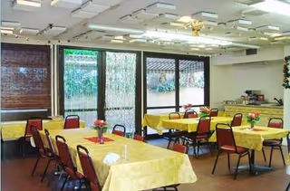 A dining room with several rectangular tables covered with yellow tablecloths, each table surrounded by red chairs. Small flower arrangements are placed on the tables. Large windows and glass doors let in natural light, and there are blinds partially covering the windows. The ceiling has multiple light fixtures and a ceiling fan.