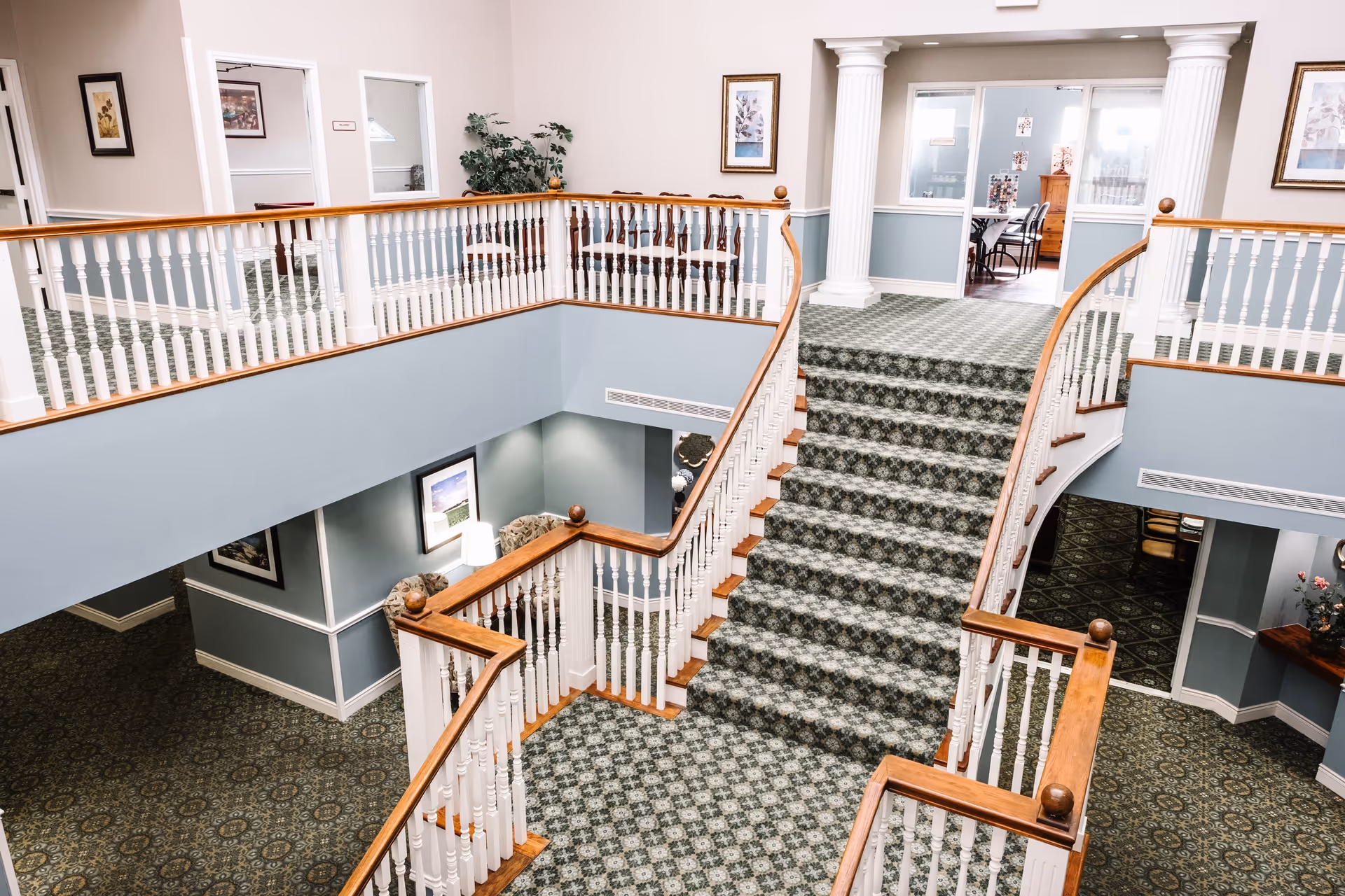 Interior view of a senior living facility with a carpeted staircase leading to an upper floor. The area features white railings with wooden handrails, blue walls, framed artwork, and decorative columns. There are seating areas and rooms visible in the background.