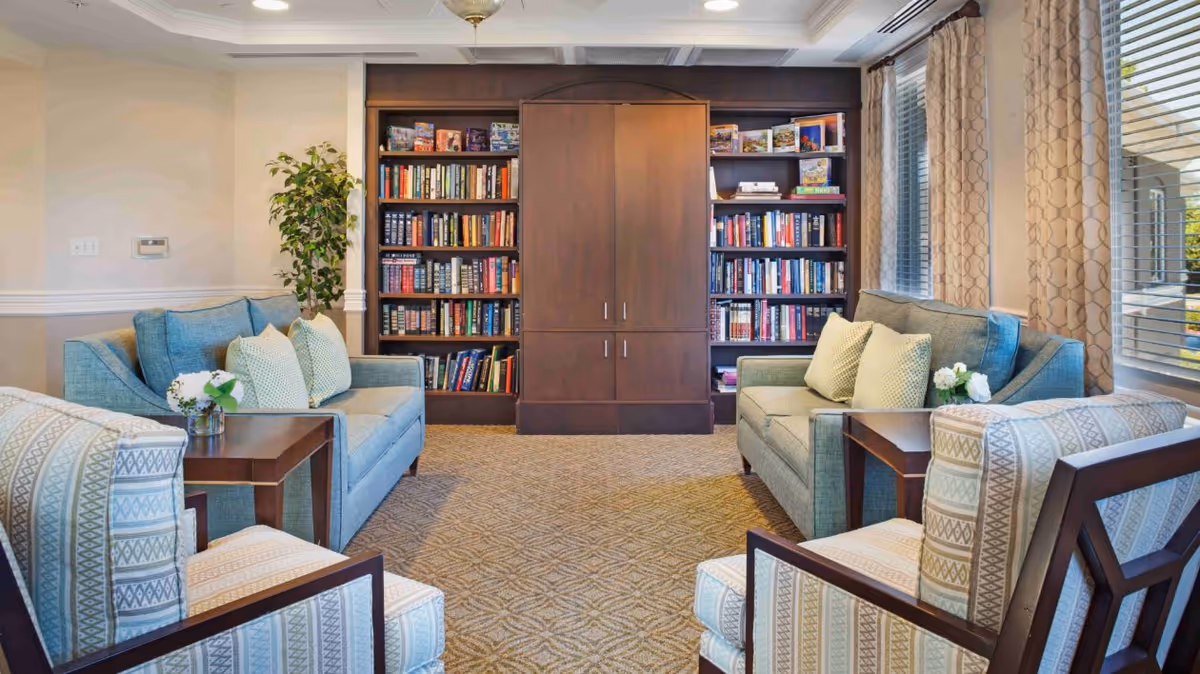A cozy living room area with two blue sofas and two patterned armchairs arranged around a central carpeted floor. There are two wooden side tables with small flower arrangements on them. A large dark wooden bookshelf filled with books is against the back wall, and large windows with patterned curtains let in natural light from the right side.