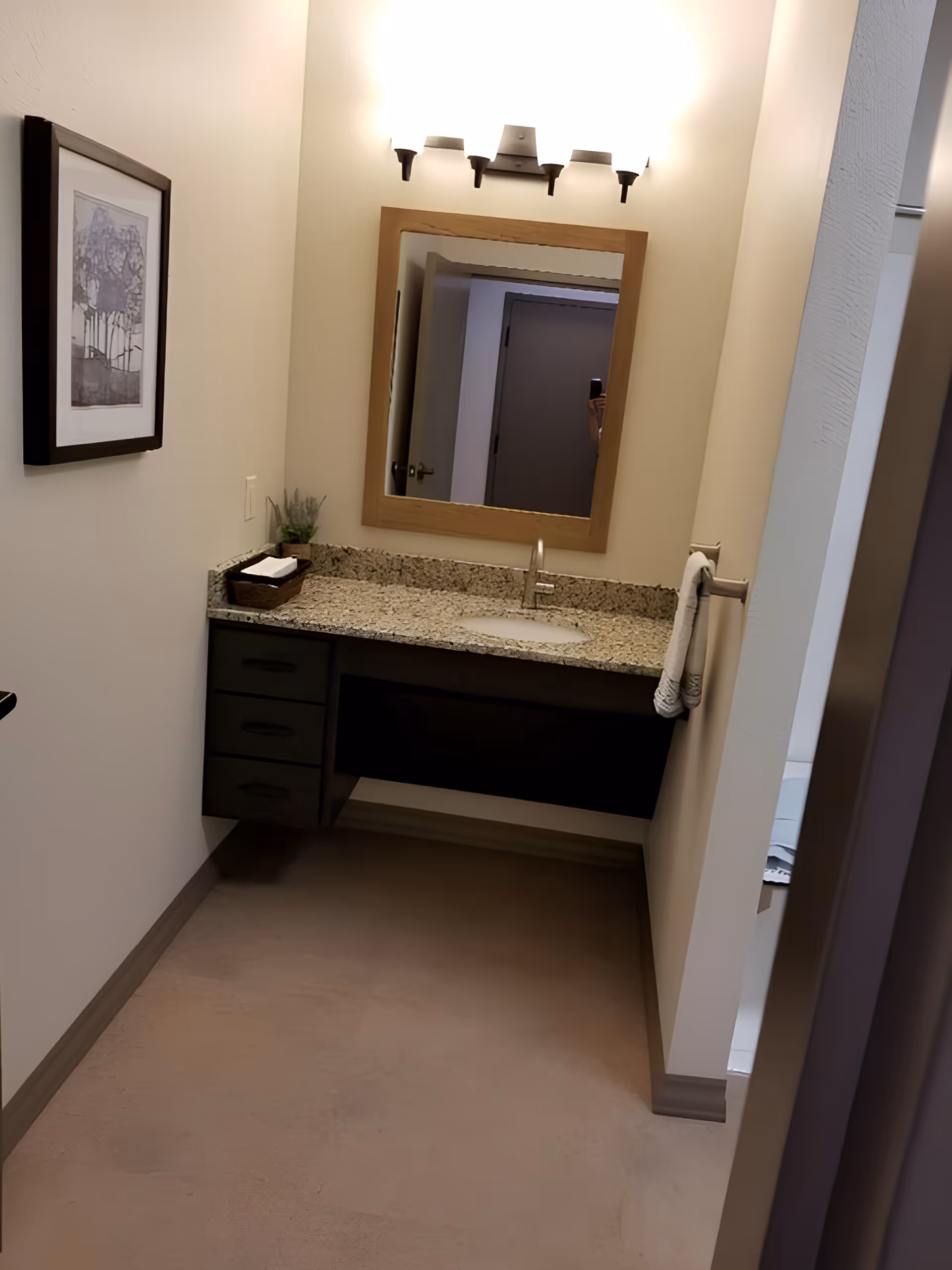 A small bathroom vanity area with a granite countertop, an under-mount sink, and a modern faucet. Above the sink is a wooden framed mirror with a three-light wall fixture. To the left of the vanity is a framed artwork on the wall, and a small basket with towels and a small plant on the countertop. A hand towel hangs on a towel ring mounted on the right wall. The floor is a smooth, light brown surface.